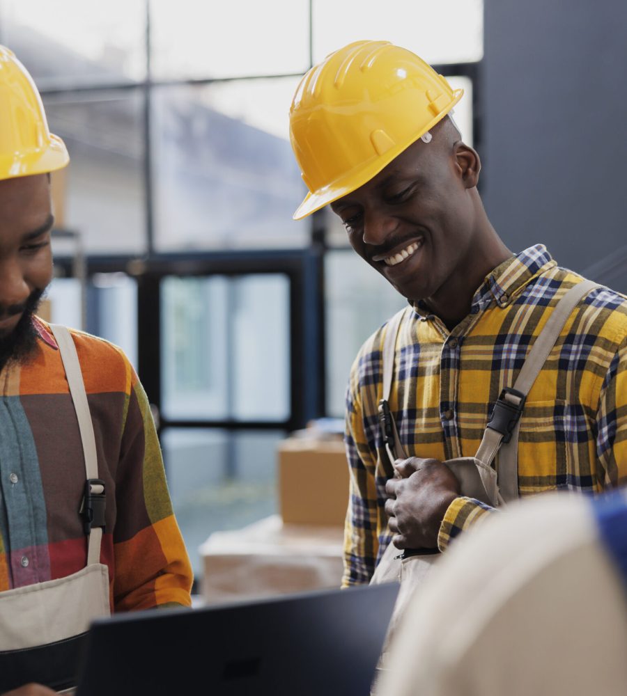 Warehouse workers wearing yellow helmets laughing and chatting at work. Smiling african american men storehouse employees in protective hard hats and overall having fun conversation